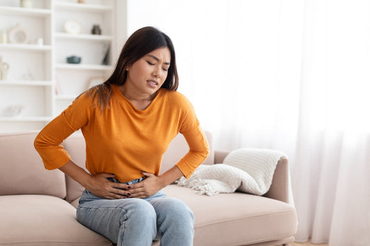 Unhappy sick pretty long-haired young asian woman wearing casual comfy outfit sitting on couch in living-room at home, touching belly, suffering from period cramps, copy space
